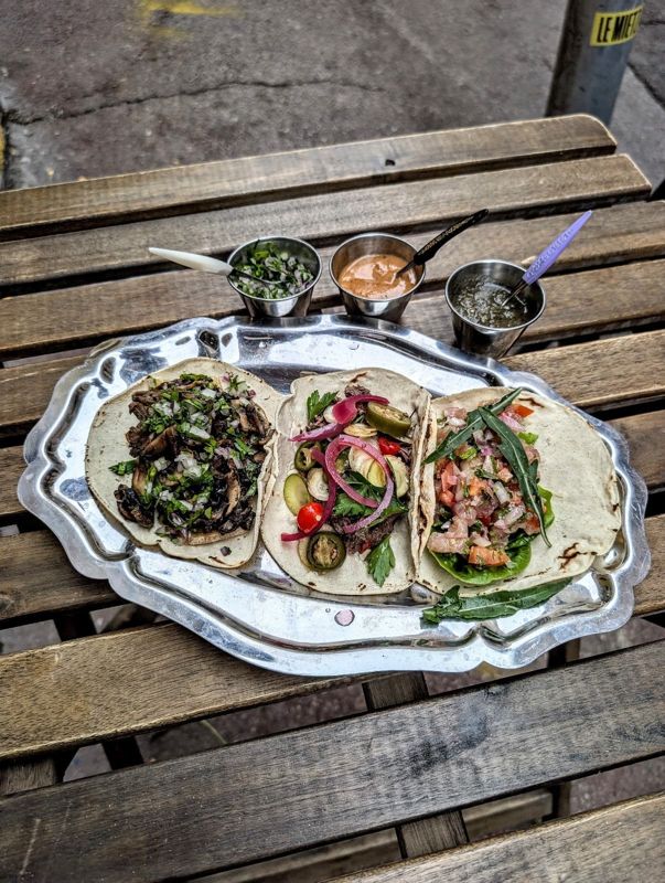 Un trio de tacos garnis sur un plateau en métal, avec trois sauces maison